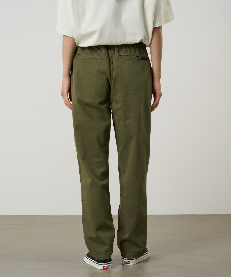 Back view of a model wearing olive green pants, a cream short-sleeve top, and black low-top sneakers, standing against a plain light gray studio background.