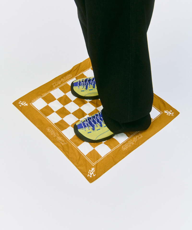Model wearing black pants standing on a yellow orange Gamicci Checkers Bandana on a light-tone background.