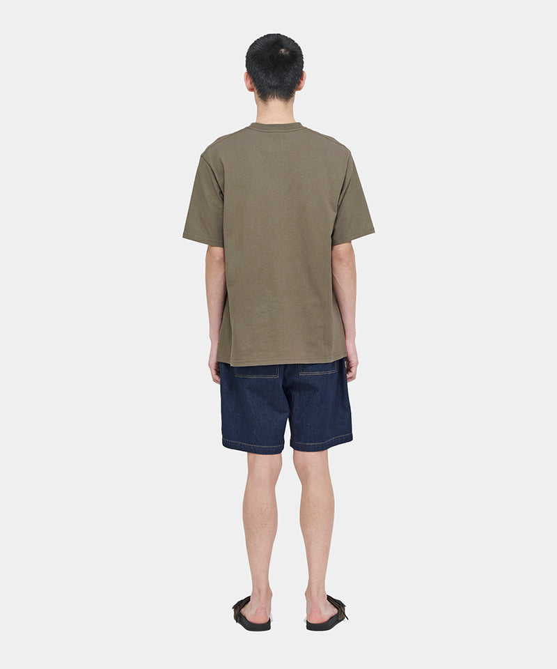 Back view of a model wearing a coyote brown Gramicci One Point Tee and blue shorts on a light-tone background.