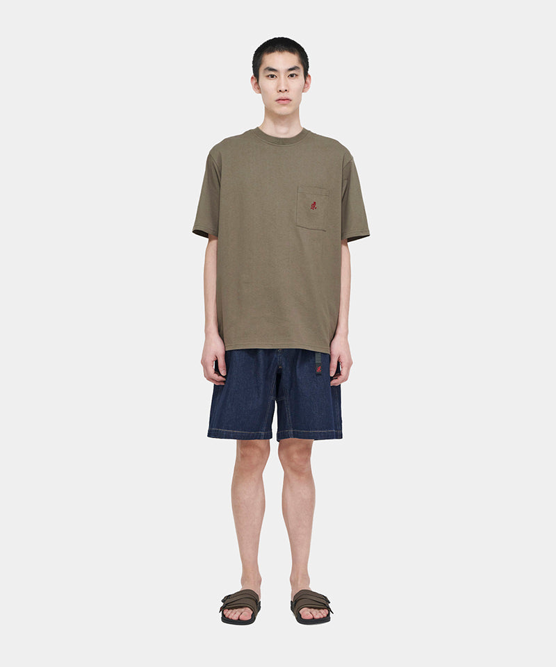 Model wearing a coyote brown Gramicci One Point Tee with a small running man graphic and blue shorts on a light-tone background.