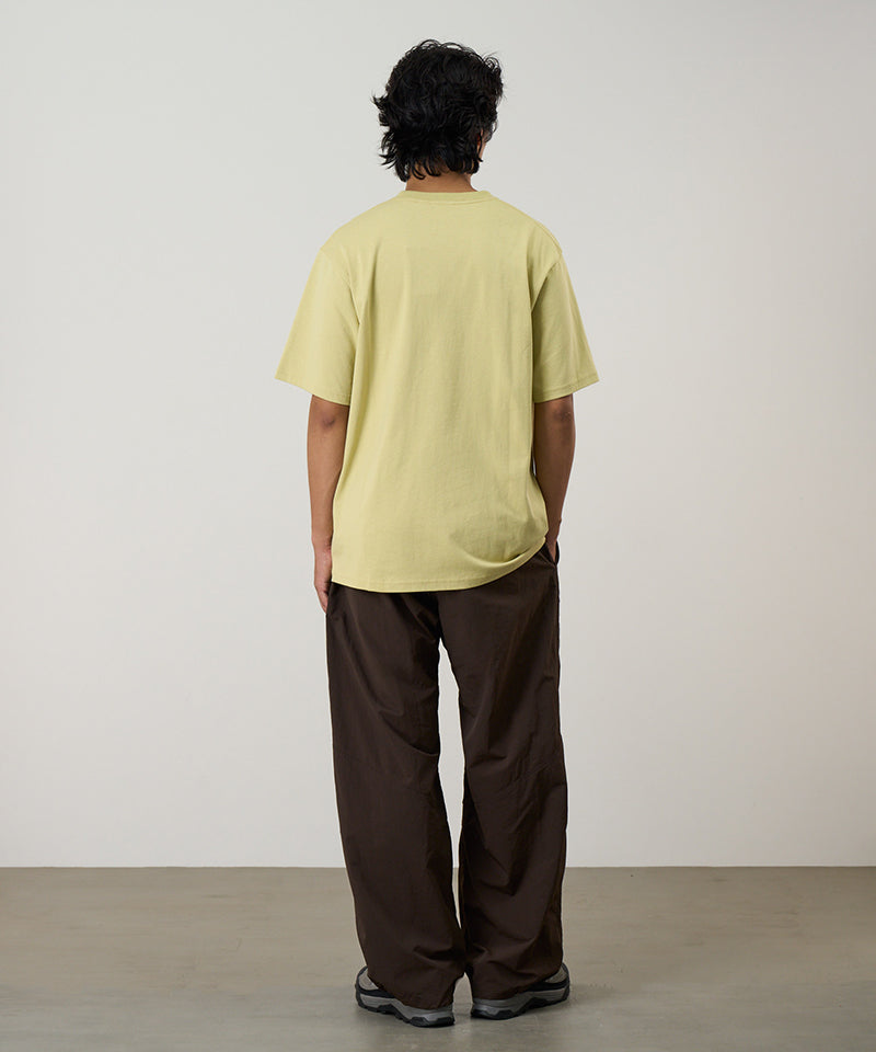 Back view of a model wearing a lime green Gramicci One Point Tee and brown pants on a light-tone background.