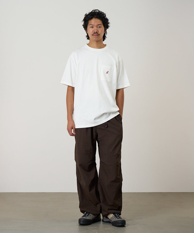 Model wearing a white Gramicci One Point Tee with a small running man graphic and brown pants on a light-tone background.