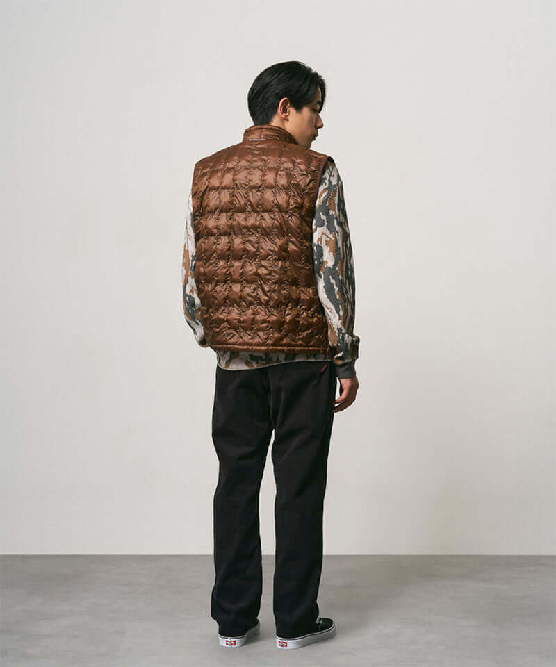 Back view of a model wearing black Gramicci Corduroy Loose Tapered Ridge Pants and a brown vest on a light-tone background.