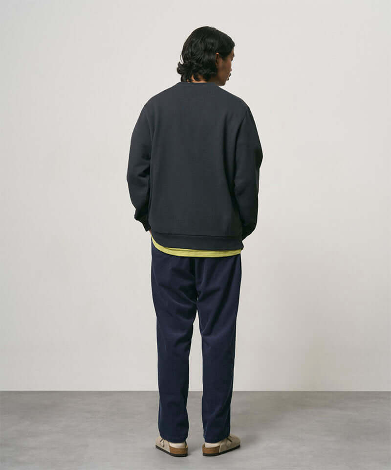 Back view of a model wearing dark navy Gramicci Corduroy Loose Tapered Ridge Pants and a black sweatshirt on a light-tone background.
