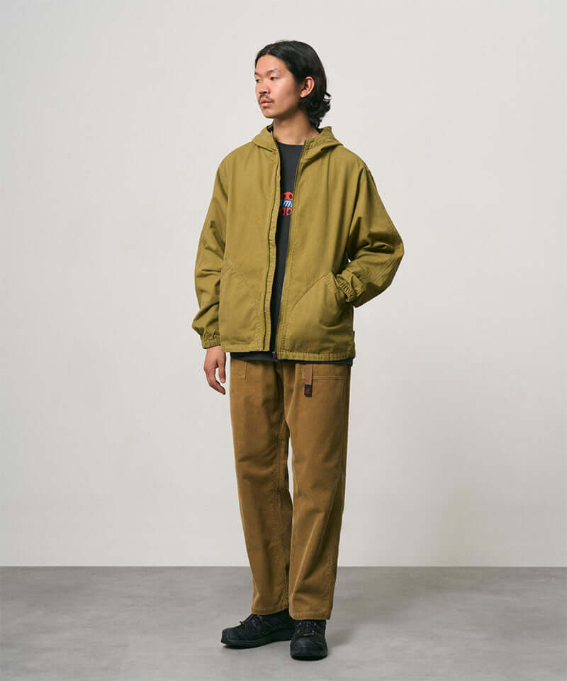 Model wearing tan Gramicci Corduroy Loose Tapered Ridge Pants and a olive colored jacket on a light-tone background.