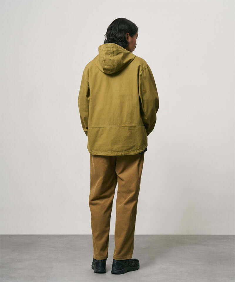Back view of a model wearing tan Gramicci Corduroy Loose Tapered Ridge Pants and a olive colored jacket on a light-tone background.