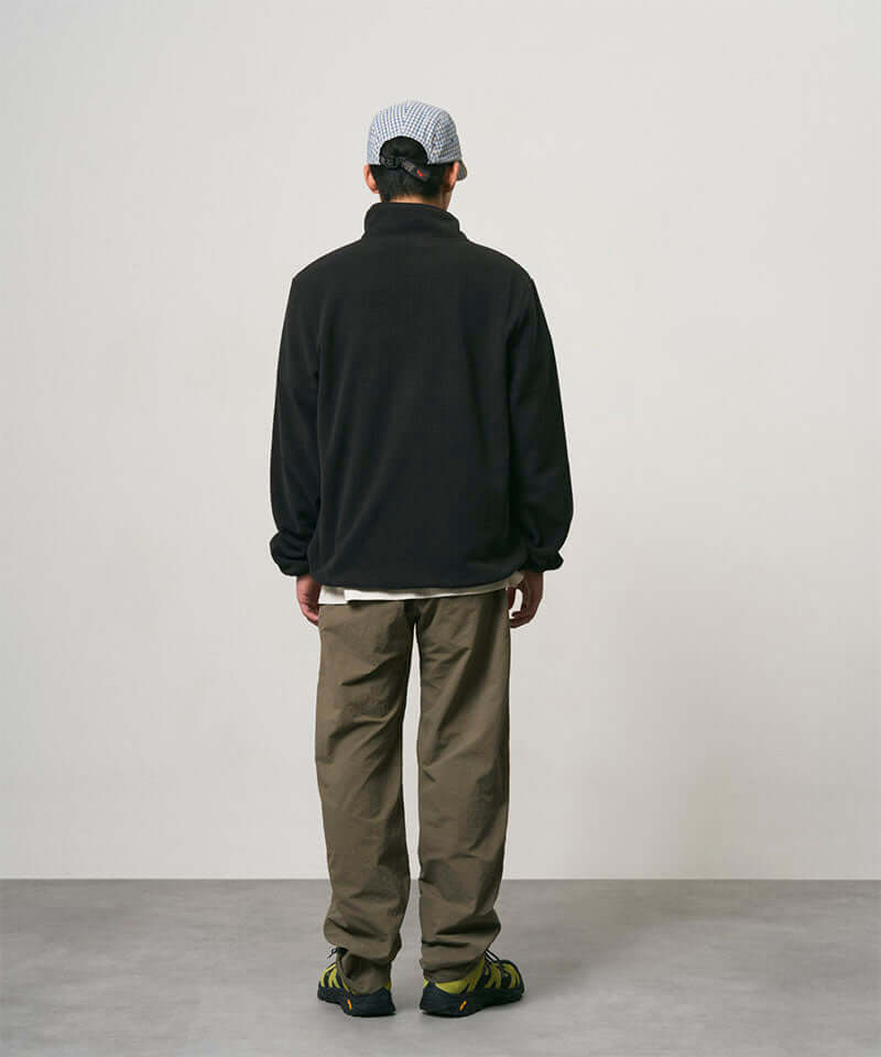 Back view of a model wearing burnt olive colored Nylon Gramicci Pants and a black jacket on a light-tone background.
