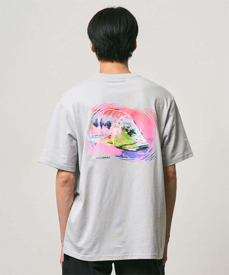 Back view of a model wearing a Gramicci mist colored Bass Tee with a graphic against a light-tone background.