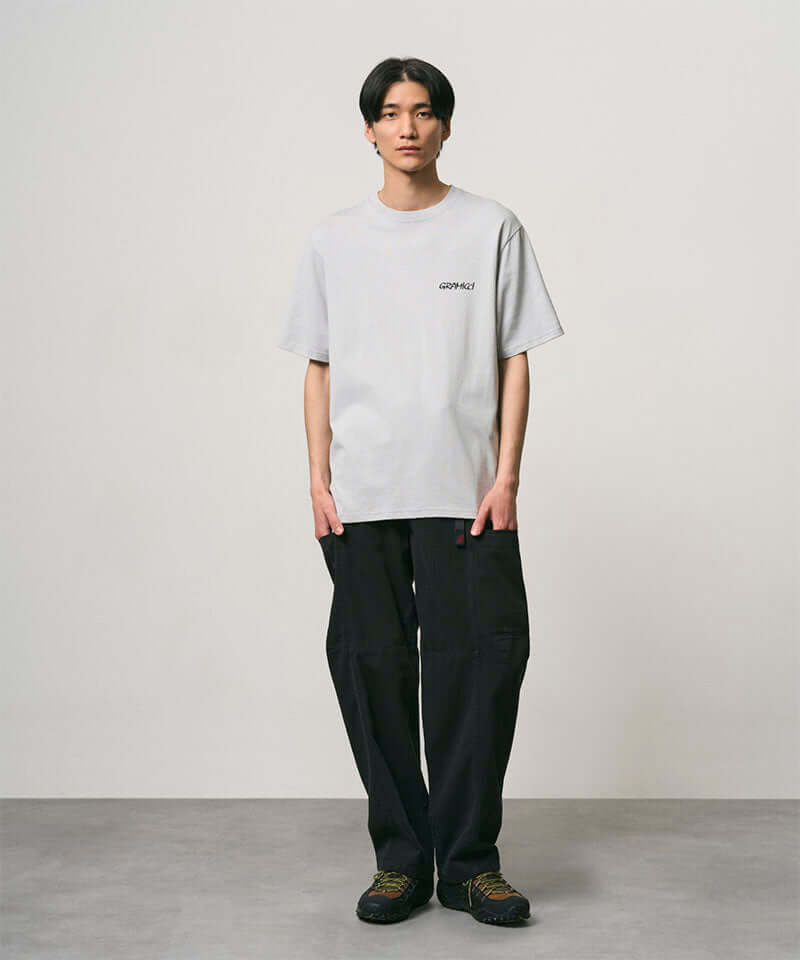 Model wearing a mist colored Gramicci Bass Tee and black pants on a light-tone background.