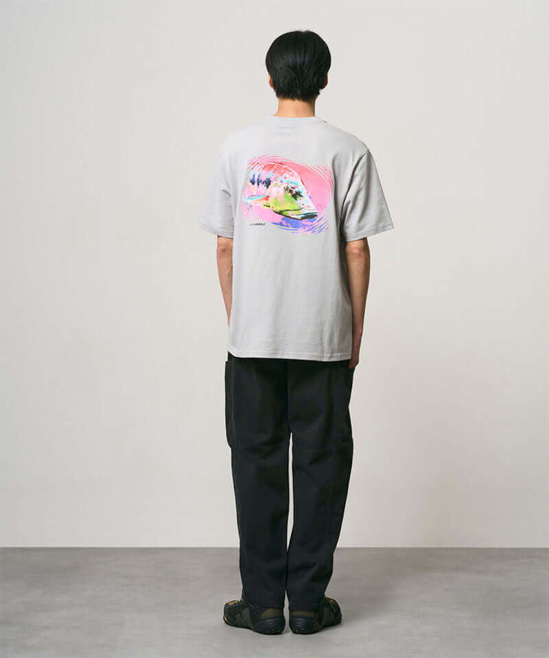 Back view of a model wearing a mist colored Gramicci Bass Tee with a graphic and black pants on a light-tone background.