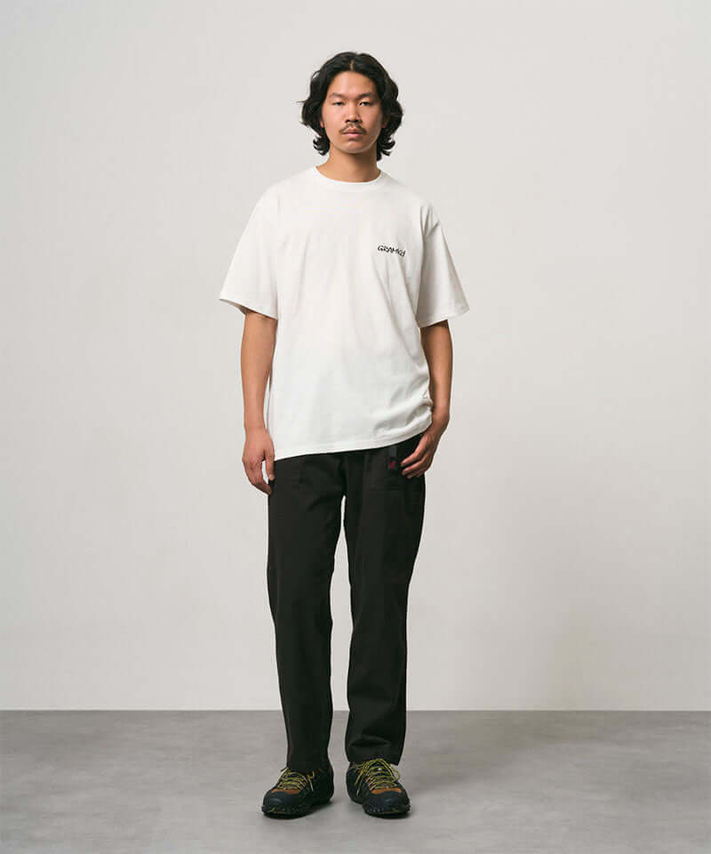 Model wearing a white Gramicci Bass Tee and black pants on a light-tone background.