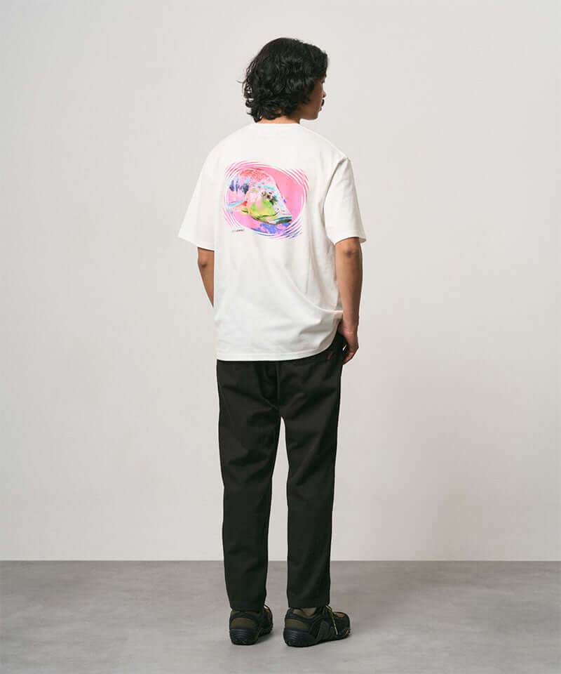 Back view of a model wearing a white Gramicci Bass Tee with a graphic and black pants on a light-tone background.
