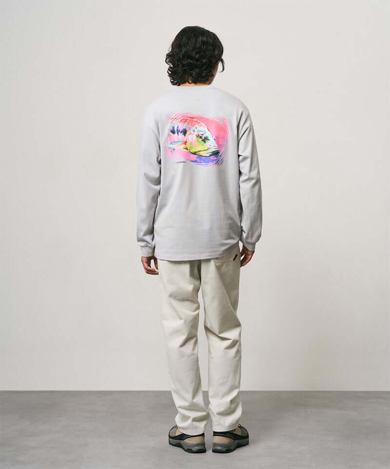 Back view of a model wearing a mist colored Gramicci Bass Long-Sleeve Tee with a graphic and off-white pants on a light-tone background.