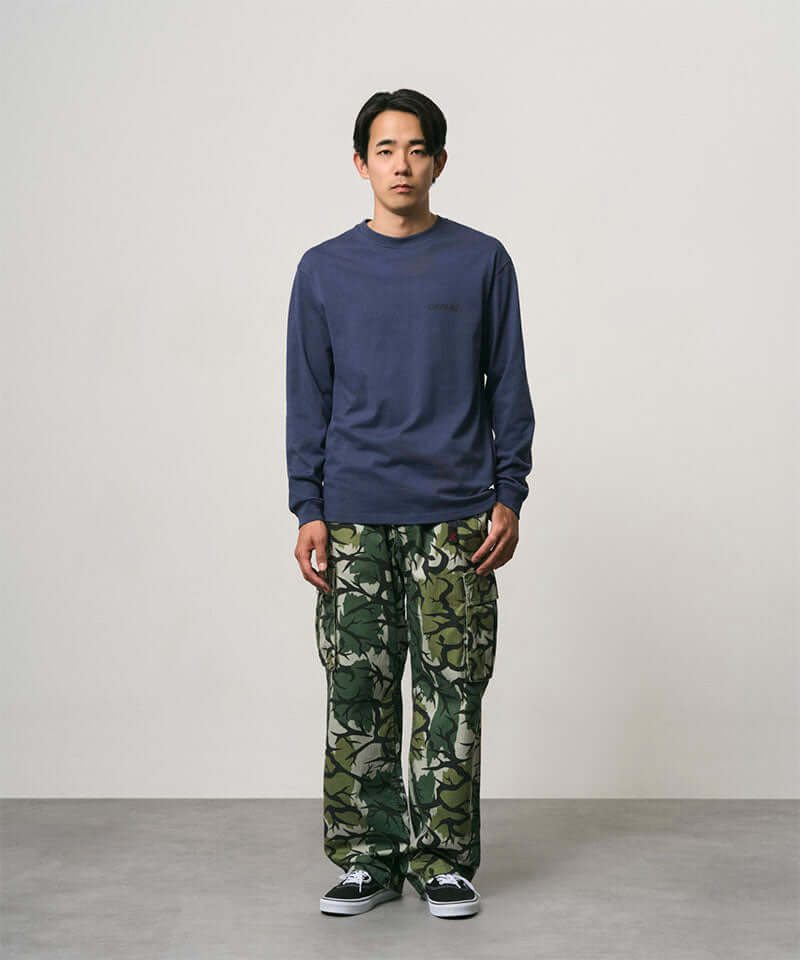 Model wearing a navy Gramicci Bass Long-Sleeve Tee and camo pants on a light-tone background.
