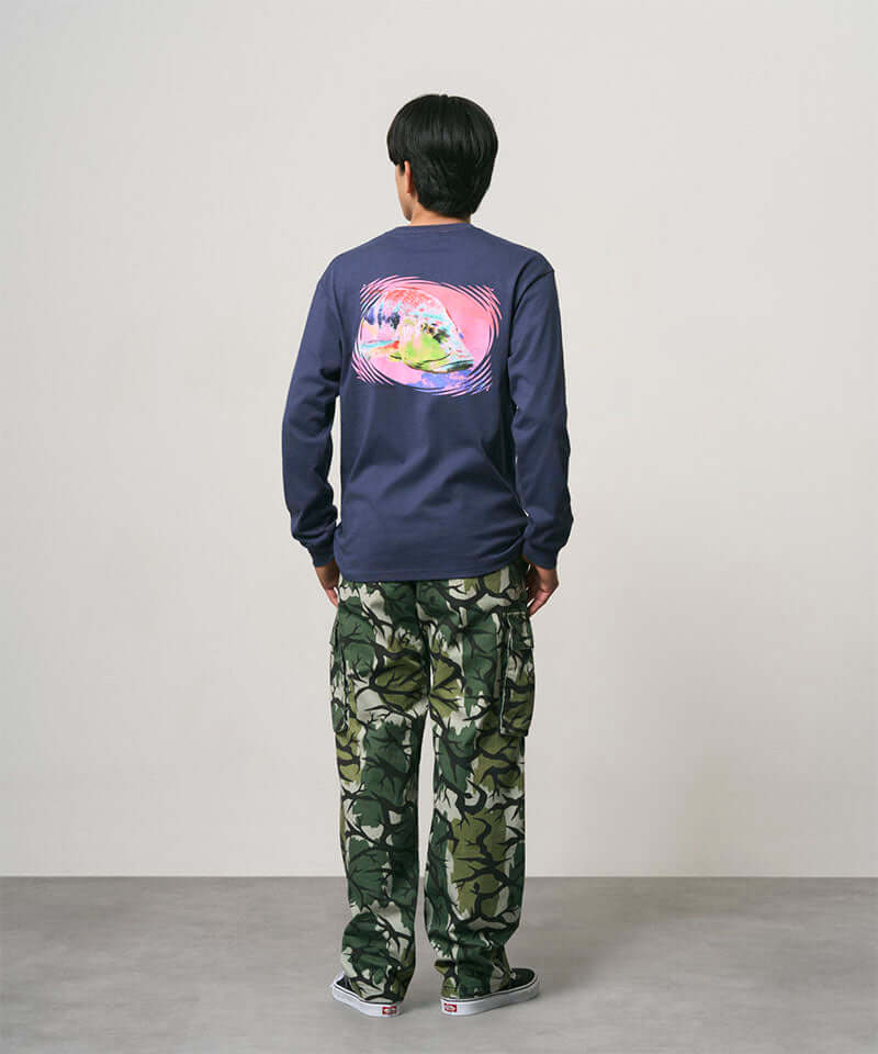 Back view of a model wearing a navy Gramicci Bass Long-Sleeve Tee with a graphic and camo pants on a light-tone background.
