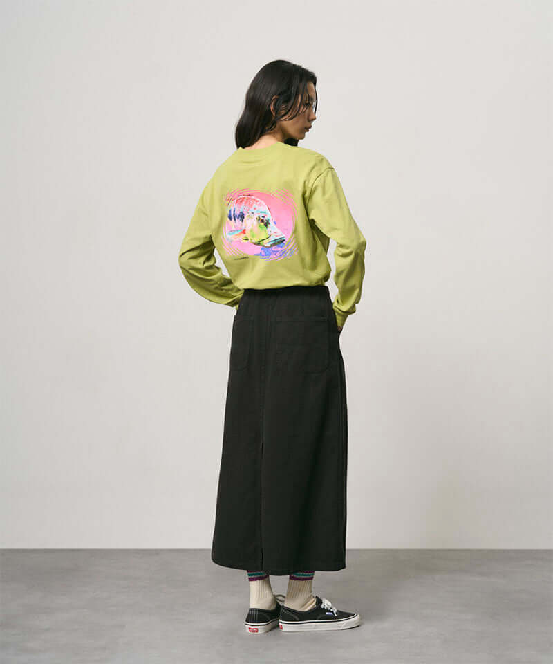 Back view of a model wearing a sweet pea colored Gramicci Bass Long-Sleeve Tee with a graphic and black skirt on a light-tone background.
