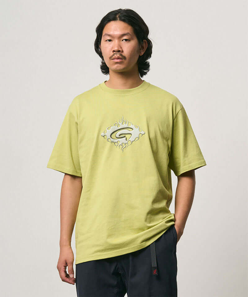 Model wearing a sweet pea colored Gramicci Flames Tee with a graphic and black pants on a light-tone background.
