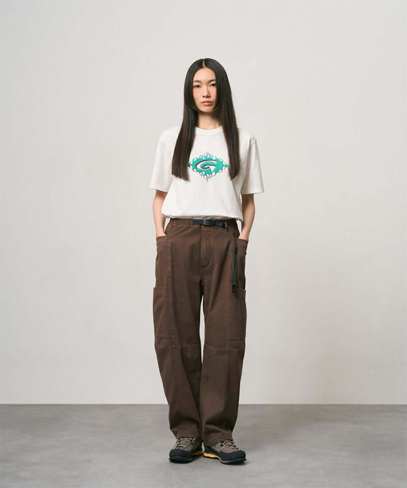 Model wearing a white Gramicci Flames Tee with a graphic and brown pants on a light-tone background.