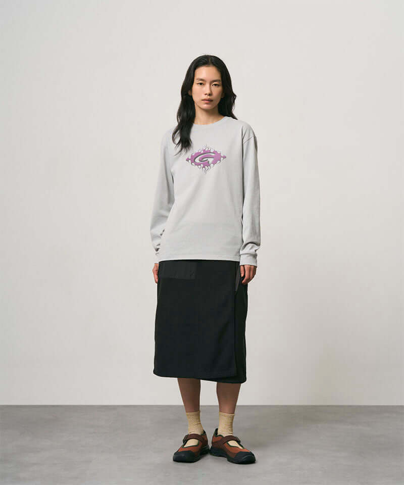 Model wearing a mist colored Gramicci Flames Long Sleeve Tee with a graphic and black skirt on a light-tone background.