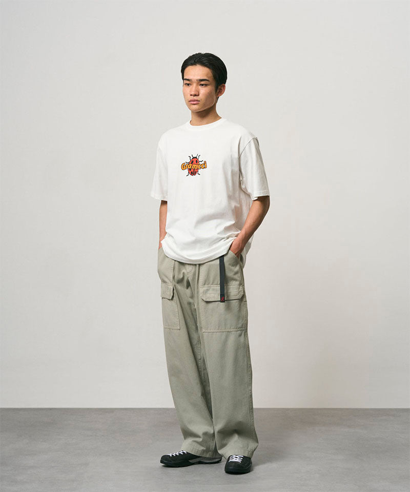 Model wearing a white Gramicci Ladybug Tee with colorful graphic and light green pants on a light-tone background.