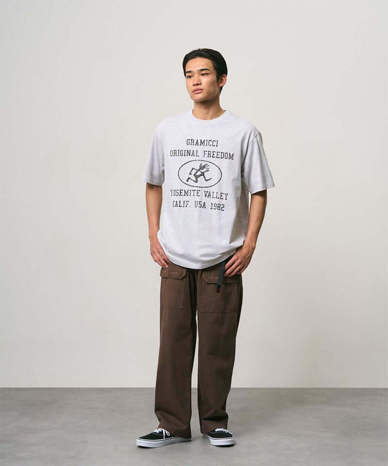Model wearing a ash heather colored Gramicci Yosemite Valley Tee with a graphic and brown pants on a light-tone background.