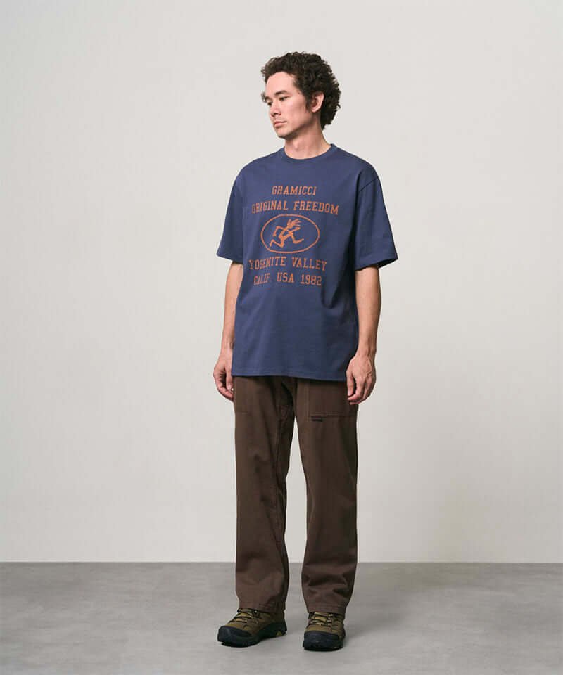 Model wearing a navy Gramicci Yosemite Valley Tee with a graphic and brown pants on a light-tone background.
