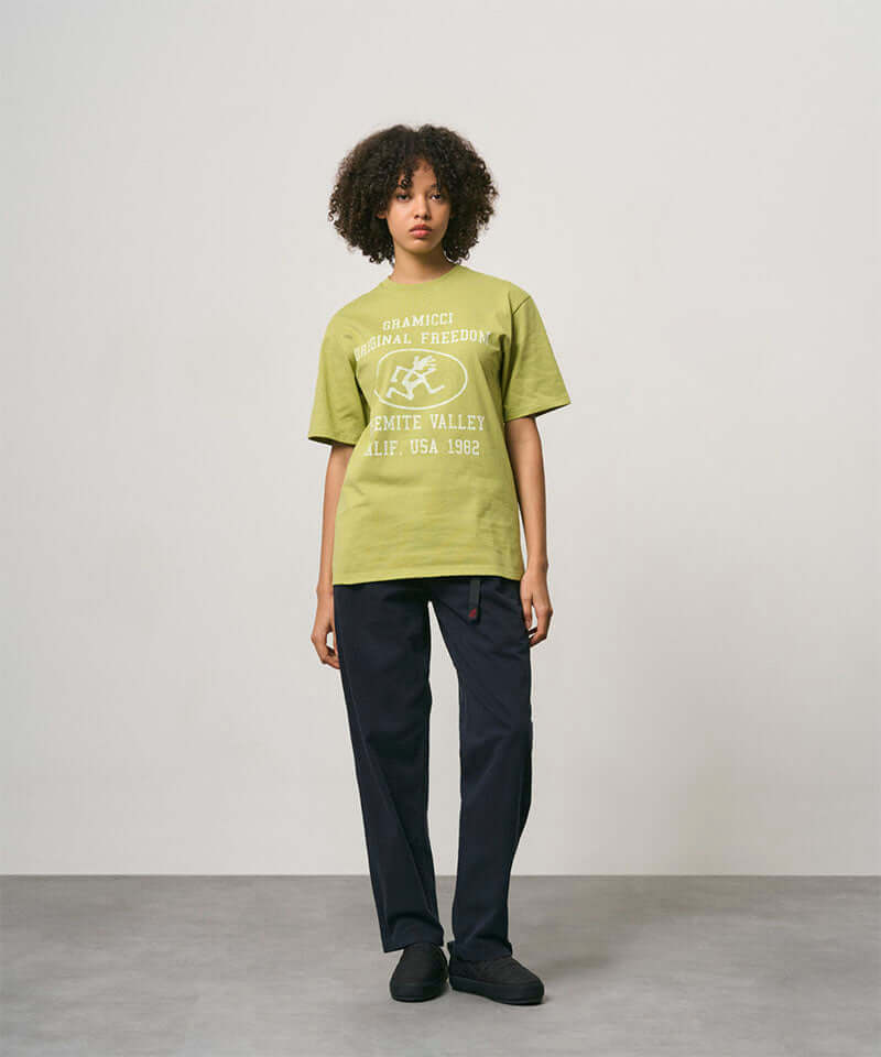Model wearing a sweet pea colored Gramicci Yosemite Valley Tee with a graphic and black pants on a light-tone background.