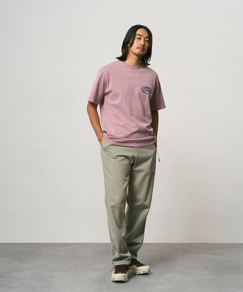 Model wearing a lilac pigment colored Gramicci GCE Tee Pigment Dyed and olive green pants on a light-tone background.