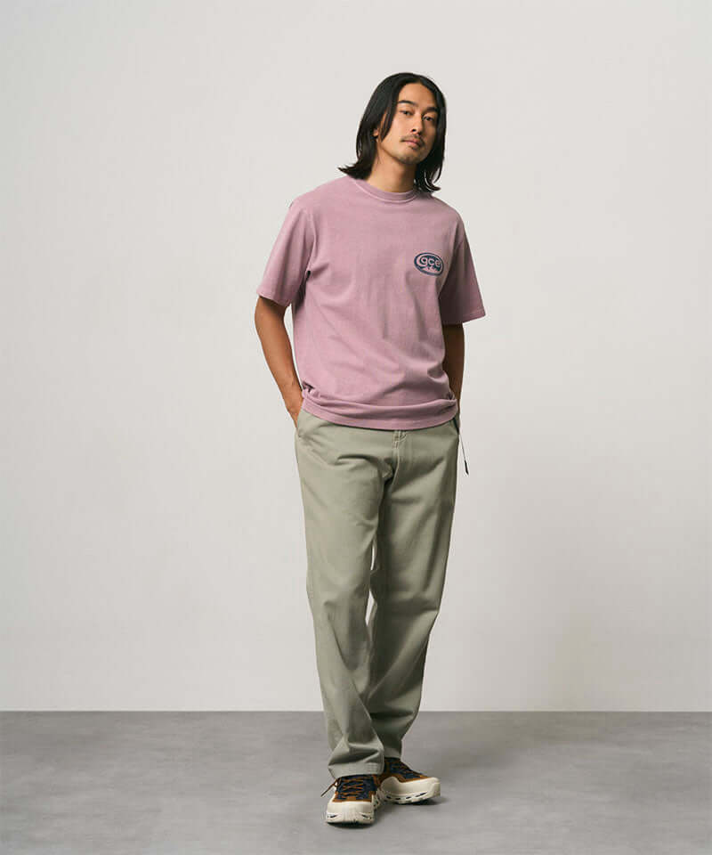 Model wearing a lilac pigment colored Gramicci GCE Tee Pigment Dyed and olive green pants on a light-tone background.