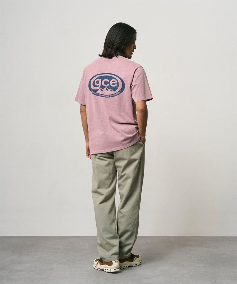 Back view of a model wearing a lilac pigment colored Gramicci GCE Tee Pigment Dyed with a graphic and olive green pants on a light-tone background.
