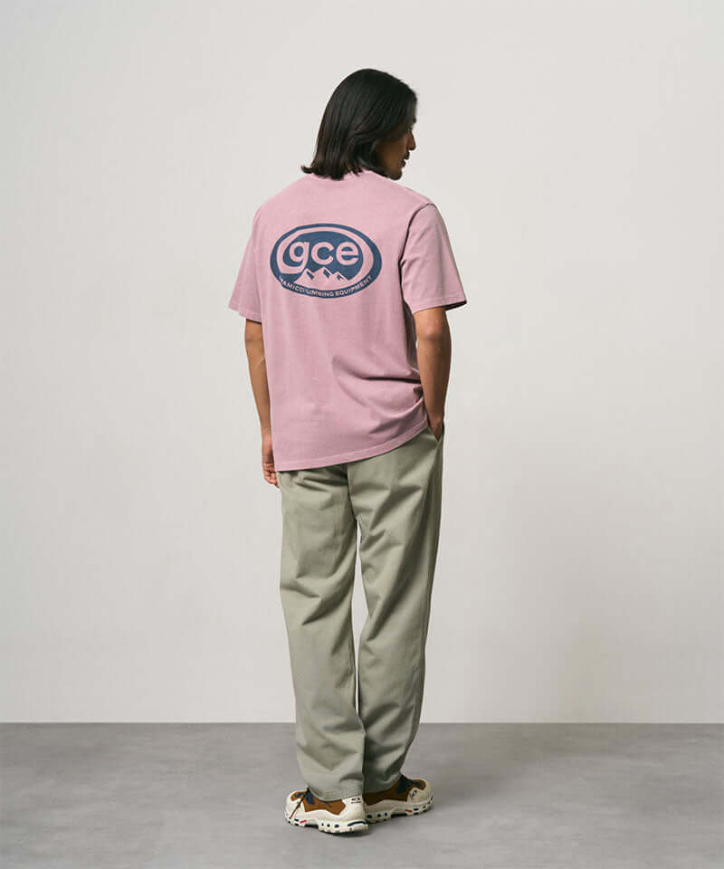 Back view of a model wearing a lilac pigment colored Gramicci GCE Tee Pigment Dyed with a graphic and olive green pants on a light-tone background.