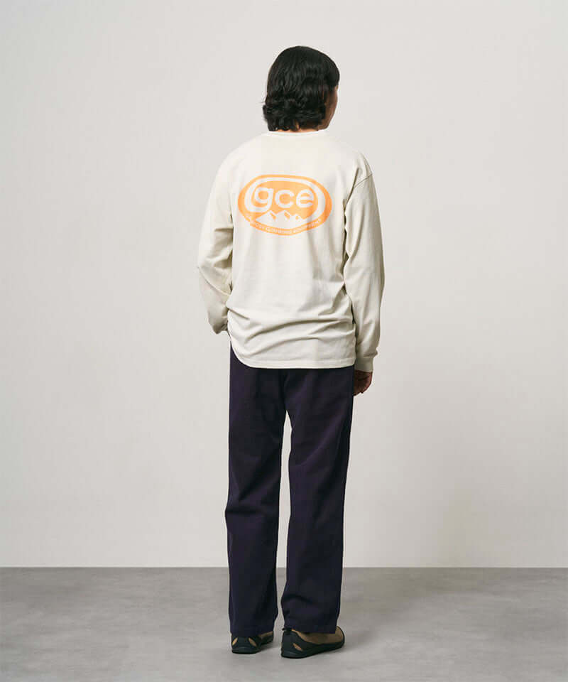 Back view of a model wearing a natural pigment colored Gramicci GCE Tee Pigment Dyed with a graphic and black pants on a light-tone background.