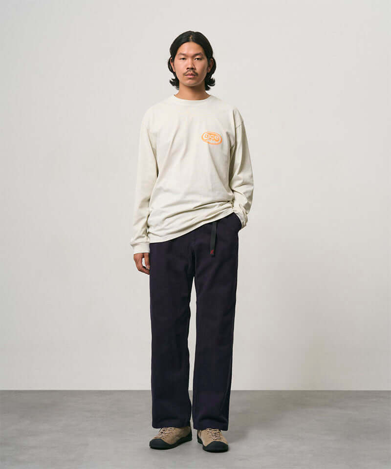 Model wearing a natural pigment colored Gramicci GCE Tee Pigment Dyed and black pants on a light-tone background.