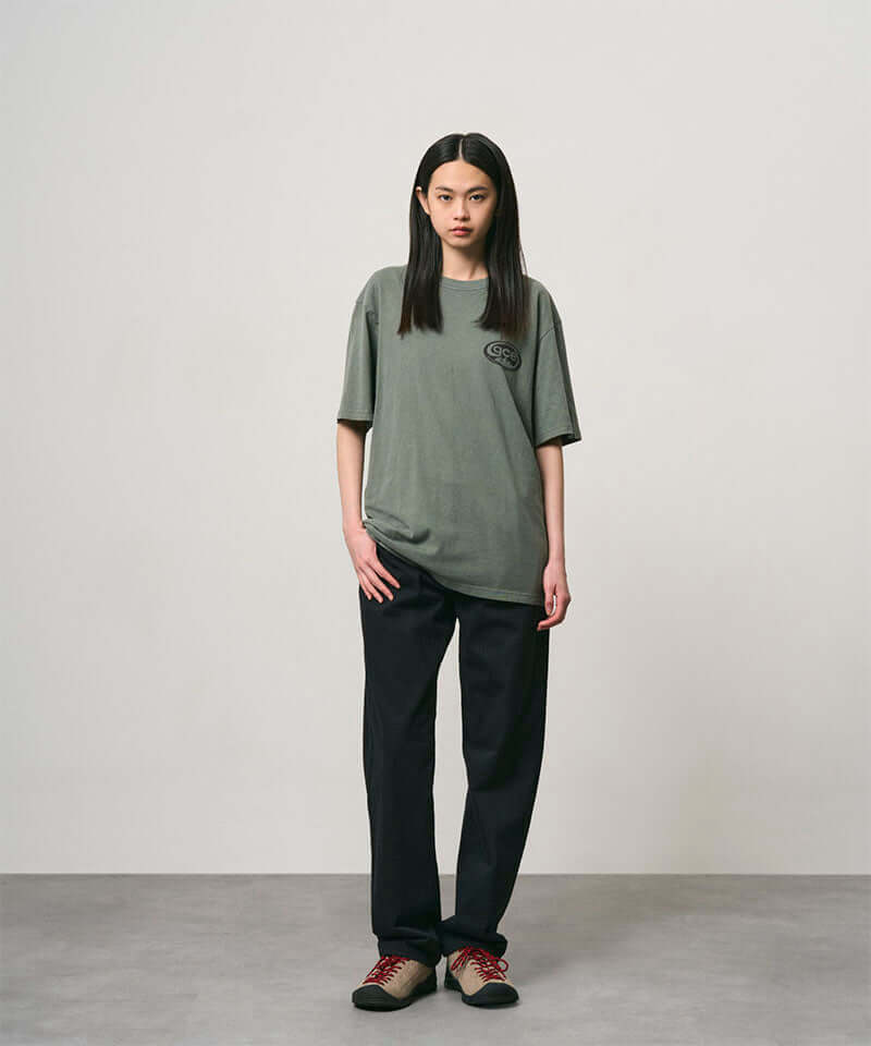 Model wearing a sage pigment colored Gramicci GCE Tee Pigment Dyed and black pants on a light-tone background.