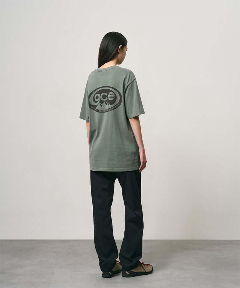 Back view of a model wearing a sage pigment colored Gramicci GCE Tee Pigment Dyed with a graphic and black pants on a light-tone background.