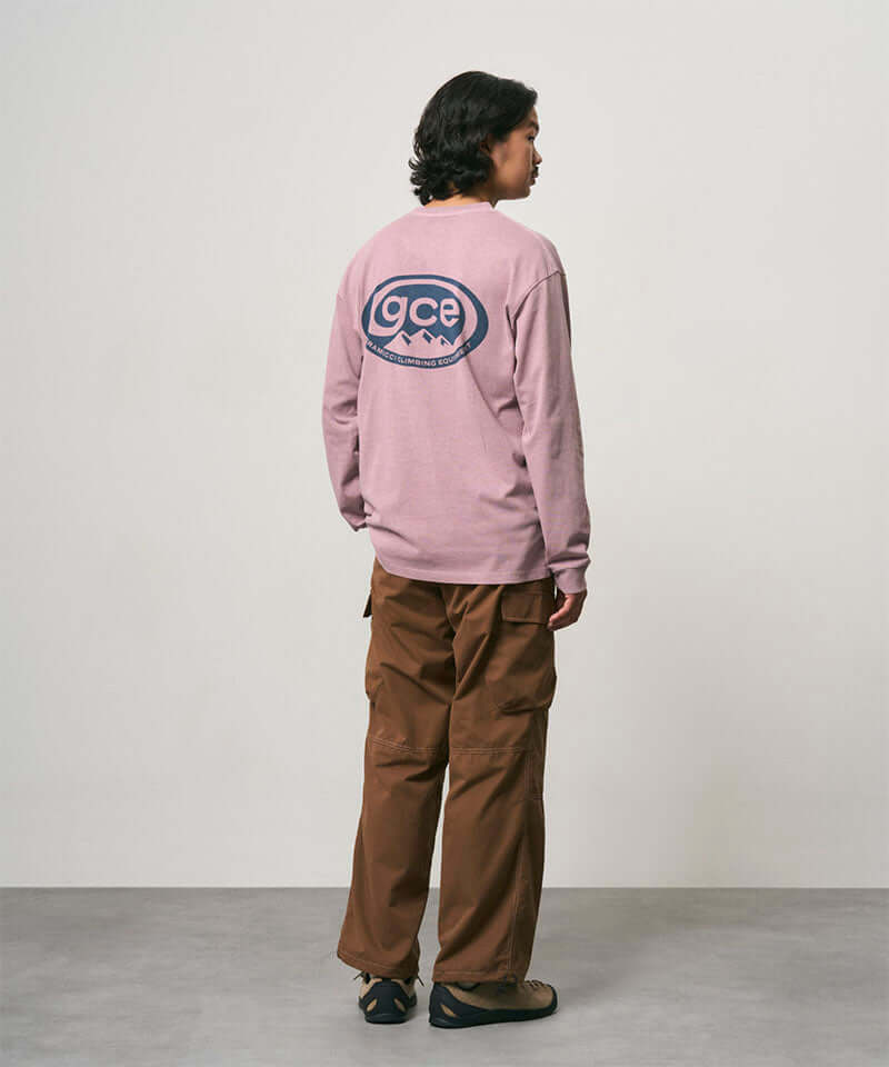 Back view of a model wearing a lilac pigment Gramicci GCE Long Sleeve Tee Pigment Dyed with a graphic and brown pants on a light-tone background.