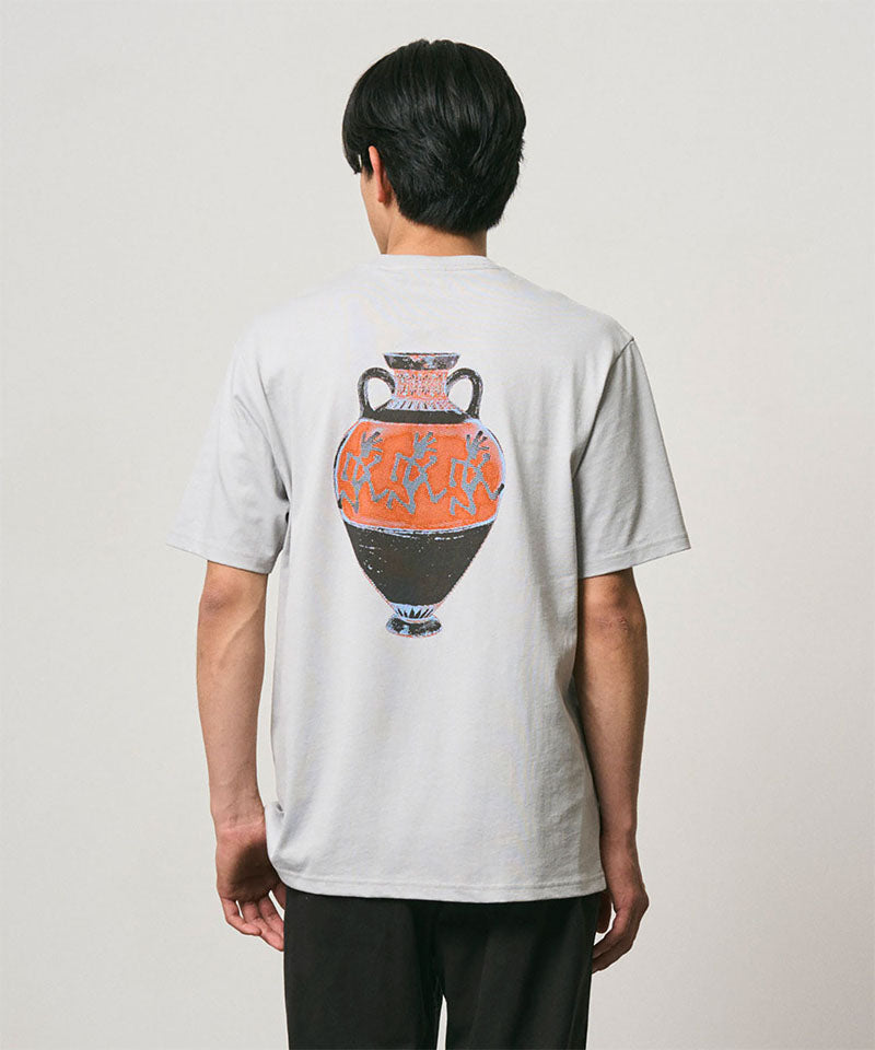 Back view of a model wearing a Gramicci mist colored Vase Tee with a graphic against a light-tone background.