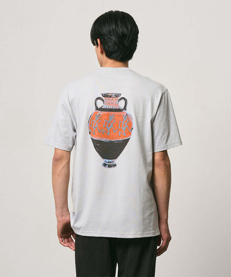 Back view of a model wearing a Gramicci mist colored Vase Tee with a graphic against a light-tone background.