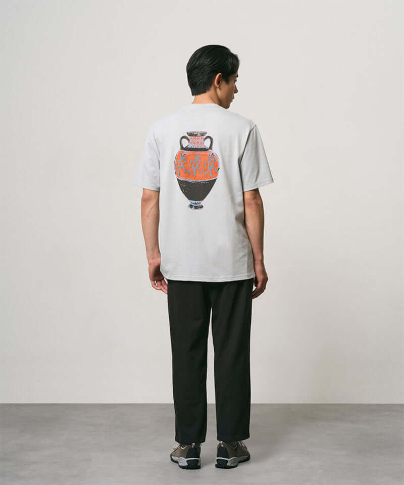 Back view of a model wearing a mist colored Gramicci Vase Tee with a graphic and black pants on a light-tone background.