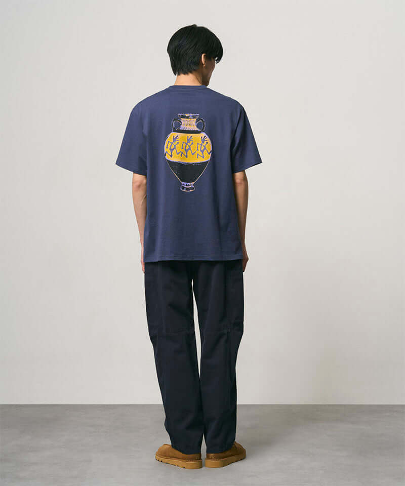 Back view of a model wearing a navy Gramicci Vase Tee with a graphic and black pants on a light-tone background.