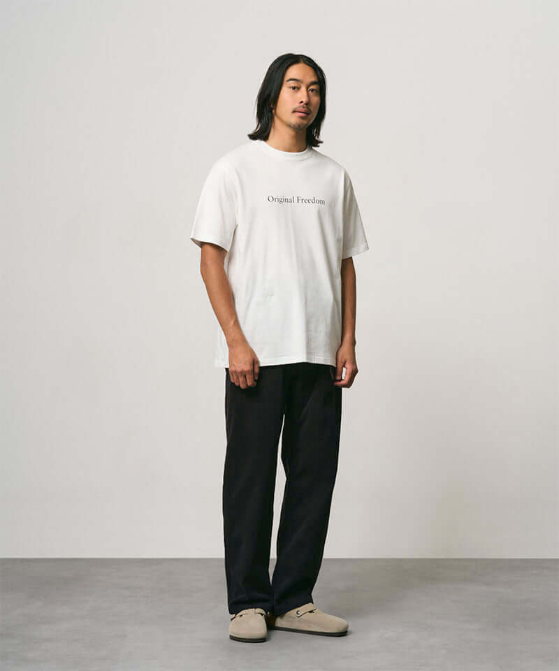Model wearing a white Gramicci Vase Tee and black pants on a light-tone background.