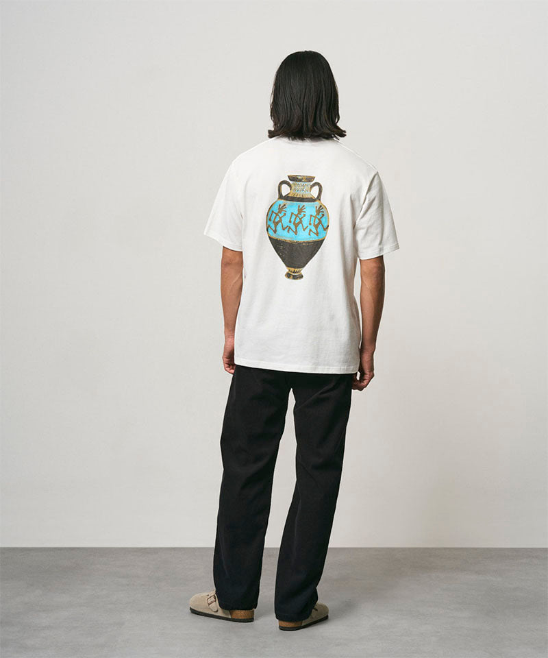Back view of a model wearing a white Gramicci Vase Tee with a graphic and black pants on a light-tone background.