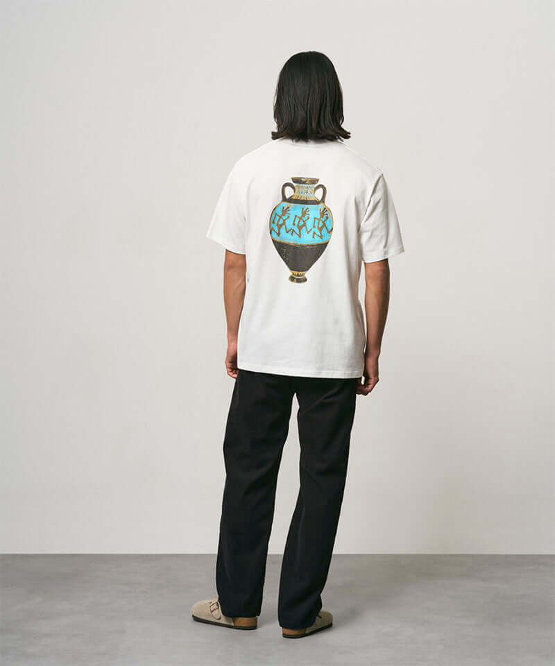 Back view of a model wearing a white Gramicci Vase Tee with a graphic and black pants on a light-tone background.