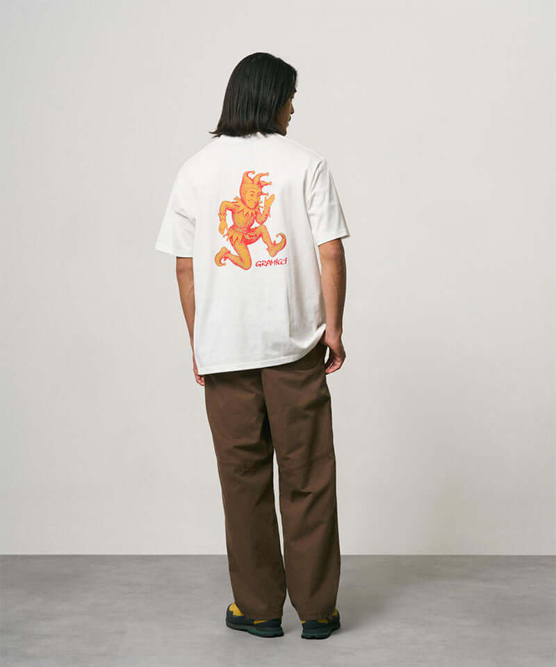 Back view of a model wearing a white Gramicci Joker Tee with an orange graphic and light green pants on a light-tone background.