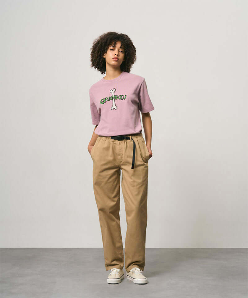 Model wearing a lailac pigment colored Gramicci Bone Tee Pigment Dyed with a graphic and beige pants on a light-tone background.