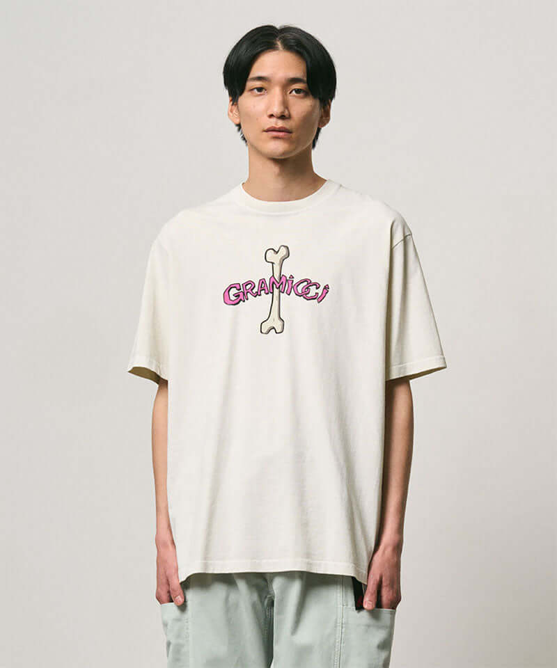 Model wearing a Gramicci natural pigment colored Bone Tee Pigment Dyed with a graphic against a light-tone background.