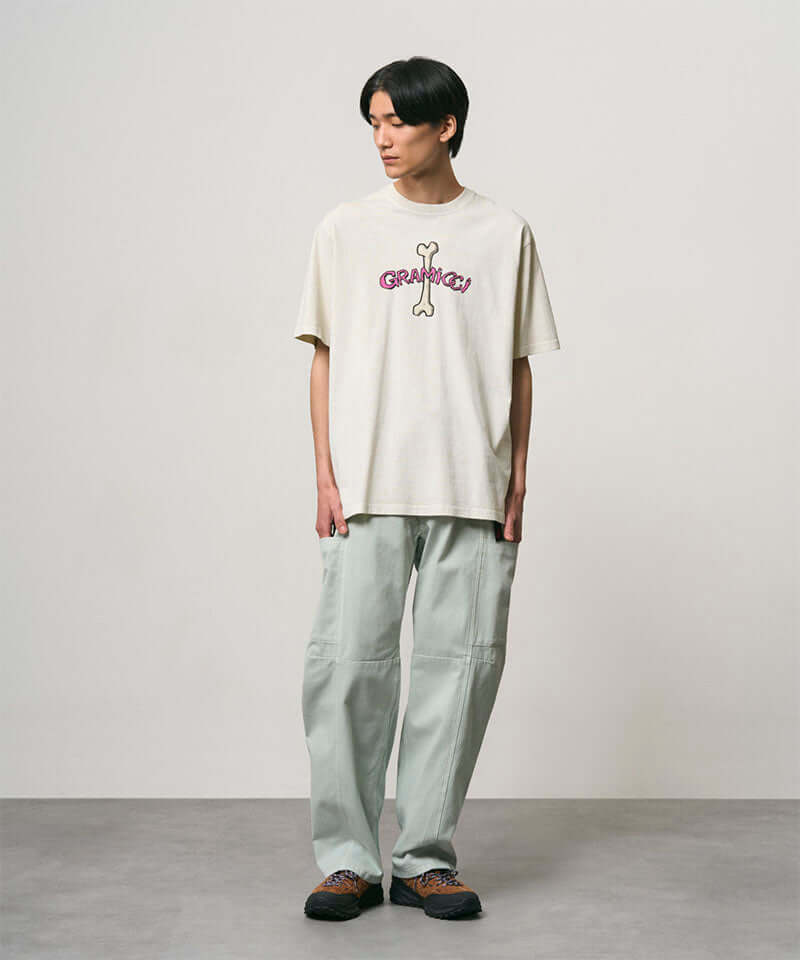 Model wearing a natural pigment colored Gramicci Bone Tee Pigment Dyed with a graphic and light green pants on a light-tone background.