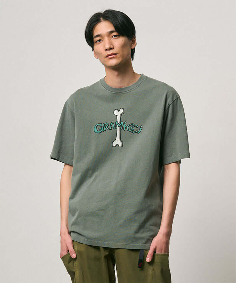 Model wearing a Gramicci sage pigment colored Bone Tee Pigment Dyed with a graphic against a light-tone background.