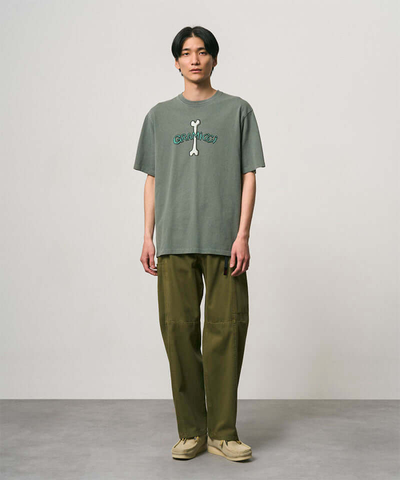 Model wearing a sage pigment colored Gramicci Bone Tee Pigment Dyed with a graphic and olive pants on a light-tone background.