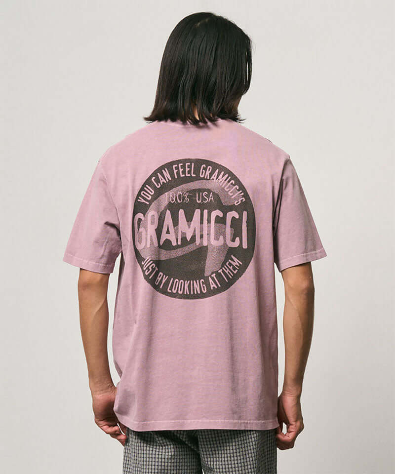 Back view of a model wearing a Gramicci lilac pigment colored Icon Tee Pigment Dyed with a graphic against a light-tone background.